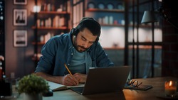 Handsome Caucasian Man With Headphones Listening Music on Laptop while Sitting in Dark Living Room in the Evening. Student Studying in Home School. Chat with Friends on Social Network.