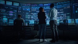 In the System Control Room Project Manager and IT Technical Engineer Have Discussion, they're surrounded by Multiple Monitors with Graphics. Big Monitor Shows Interactive Server Blockchain Info.