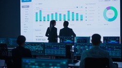 Project Leader, Chief Executive Discuss Data Shown on Big Display. Screens Show Infographics, Charts, Finance Analysis, Stock Market, Growth.Telecommunications Control Room with Working Professionals