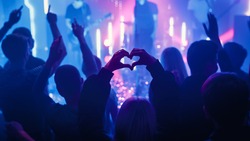 Person is Making a Heart Sign Gesture and Holding Hands Up at a Performance. Rock Band Playing a Song at a Concert in a Night Club on Stage with Bright Colorful Strobing Lights.