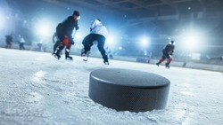 Close-up Shot with Focus on a 3D Hockey Puck on Ice Hockey Rink Arena. In the Background Blurred Professional Players From Different Teams Trying to Get the Puck. Dutch Angle Shot.