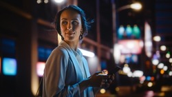 Beautiful Young Woman Using Smartphone Walking Through Night City Street Full of Neon Light. Smiling Female Using Mobile Phone for Online Shopping, Social Media Posting.
