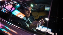 Handsome Businessman in a Suit Commuting from Office in a Backseat of His Luxury Car at Night. Entrepreneur Using Smartphone while in Transfer Taxi in Urban City Street with Working Neon Signs.