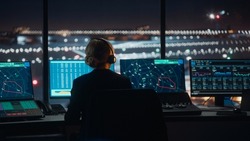 Female Air Traffic Controller with Headset Talk on a Call in Airport Tower at Night. Office Room is Full of Desktop Computer Displays with Navigation Screens, Airplane Flight Radar Data for the Team.