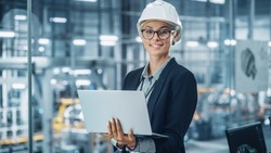 Portrait of a Happy Young Beautiful Female Engineer Wearing White Hard Hat, Using Laptop Computer in Office at Car Assembly Plant. Industrial Specialist Working on Vehicle Design in Modern Facility.
