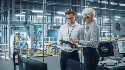 Male Engineer and Female Industrial Product Designer Discuss Work while Standing in Office at a Car Assembly Plant. Industrial Specialists Working on Vehicle Design in Technological Factory.