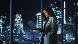 Big City Modern Office at Night: Successful Young Businesswoman Standing and Using Laptop. Beautiful Female Digital Entrepreneur Thinking of Investment Strategy for e-Commerce Project.