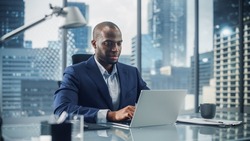 Successful Black Businessman in Tailored Suit Working on Laptop Computer on Top Floor Office Overlooking Big City. Professional CEO Managing Environmental, Social and Corporate Governance