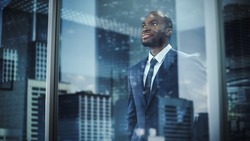 Thoughtful African-American Businessman in a Perfect Tailored Suit Standing in His Office Looking out of the Window on Big City. Successful Investment Manager Planning Strategy. Outside Shot
