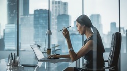 Successful Businesswoman in Stylish Dress Sitting at a Desk in Modern Office, Using Laptop Computer, Next to Window with Big City View. Successful Finance Manager Planning Work Projects.