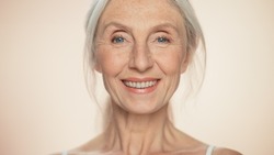 Portrait of Senior Woman Looking at Camera, Smiling. Elderly Lady with Natural Grey Hair, Blue Eyes. Graceful of Old Age Concept for Skincare, Beauty, Cosmetics Product with Isolated Beige Background