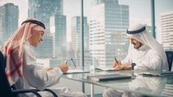 Two Happy Emirati Businessmen in White Traditional Kandura Sitting in Office and Signing Contract. Business Partners Sign Lucrative Investment. Saudi, Emirati, Arab Businessman Concept.