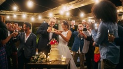 Beautiful Bride and Groom Celebrate Wedding at an Evening Reception Party with Multiethnic Friends. Married Couple Standing at a Dinner Table, Kiss and Cut Wedding Cake.