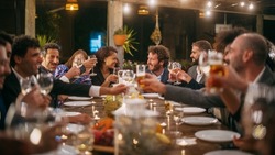Big Dinner Party with a Small Crowd of Multiethnic Diverse Friends Celebrating at a Restaurant. Beautiful Happy Hosts Propose a Toast and Raise Wine Glasses while Sitting at a Table in the Evening.