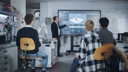 Teacher Pointing at Big Screen and Holding Lesson while Robotic Arm Moving Nearby. Diverse Group of Young Engineers Sitting at University and Asking. Computer Science Education Concept
