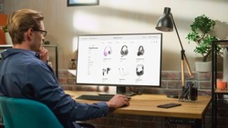 At Home Office: Man Using Desktop Computer, Online Shopping for Electronics, Wireless Hi-Fi Headphones. e-Commerce Concept of Purchasing, Buying, Ordering Tech Devices on Website. Over Shoulder View