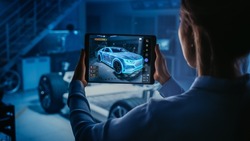 Automotive Engineer Working on Electric Car Chassis Platform, Using Tablet with Augmented Reality 3D Software. Innovative Facility Concept: Vehicle Frame with Wheels Becomes a VFX Virtual Model.