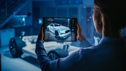 Automotive Engineer Working on Electric Car Chassis Platform, Using Tablet Computer with Augmented Reality 3D Software. Innovative Facility: Vehicle Frame with Wheels Becomes a VFX Model.