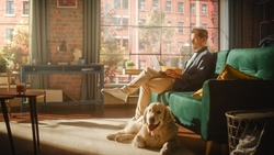 Handsome Young Man Using Laptop, Working from Home Living Room with Big Windows. Male Sitting on Couch, Using Computer, Golden Retriever Dog Sitting Next to Him and Wanting to Play or Go For a Walk.