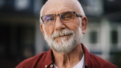 Close Up Portrait of a Cheerful Senior Man with Gray Hair Wearing Glasses Standing Outdoors in Front of a Residential Area Home. Retired Adult Man Looking at Camera and Smiling.