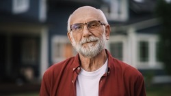 Portrait of a Happy Senior Man with Gray Hair Wearing Glasses and a Red Shirt Standing Outside in Front of a Suburbs Area House. Old Adult Man Posing and Looking at Camera and Smiling.