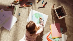 Top View: Little Girl Drawing Our Beautiful Green Planet Earth. Child Having Fun at Home on the Floor, Imagining Our Planet as a Happy Place with Clean, Sustainable Living. Cozy Sunny Day.