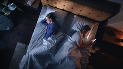 Top View Bedroom Apartment: Man Uses Smartphone in Bed at Night When His Female Partner Trying to Fall Asleep. Couple After Fight, Argument. Addictive World of Social Media, Doom Scrolling, Fake News.