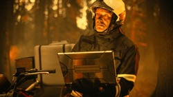 Portrait of a Handsome Fireman in Safety Gear Using Heavy-Duty Laptop Computer, Reporting on a Situation with a Dangerous Wildland Fire in a Forest. Hard Day at Work for a Brave Firefighter Volunteer.