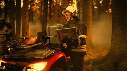 Brave Skillful Firefighter Standing Next to an ATV, Using Laptop Computer in Forest with Raging Brushfire. Superintendent or Squad Leader Making Sure Emergency Situation is Under Control.