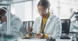 Diverse Team of Multiethnic Young Scientists Passing Internship in a Modern High Tech Laboratory. African Female Working with Soldering Iron Wearing Safety Goggles and White Lab Coat