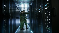 In Data Center Two Military Men Work with Open Server Rack Cabinet. One Holds Military Edition Laptop.