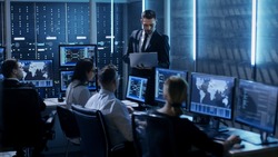 Professional IT Engineers Working in System Control Center Full of Monitors and Servers. Supervisor Holds Laptop and Holds a Briefing. Possibly Government Agency Conducts Investigation. 