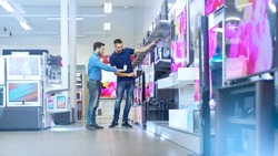 In the Electronics Store Professional Consultant Shows Latest TV's to a Young Man, They Talk about Specifications and What Model is Best for Young Man's Home. Store is Bright and Has Latest Models.