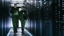 Two Military Men Walking in Data Center Corridor. One Uses Tablet Computer, They Have Discussion. Rows of Working Data Servers by their Sides.