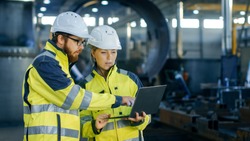 Male and Female Industrial Engineers in Hard Hats Discuss New Project while Using Laptop. They Make Showing Gestures.They Work in a Heavy Industry Manufacturing Factory.