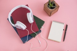 white headphones lie on pile of books on pink background. concept: audiobook