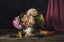 Still life with gentle hydrangea, a cup of tea and elderberry jam and a basket of elderberries on a wooden table