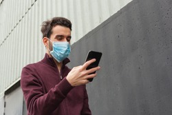 White bearded adult man using smartphone while wearing surgical mask on an industrial wall. Health, epidemics, social media, communication and lifestyle stills with copy space.