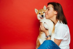 Cute brunette woman in white t shirt and jeans holding and embracing Shiba Inu dog, has eyes closed with pleasure, isolated on red background. Love to the animals, pets concept