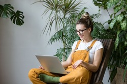 Woman gardener in glasses wearing overalls, sitting on wooden chair in greenhouse, using laptop after work, talks with her friend about coronavirus and stay home during online video call. Working home
