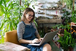 Woman gardener in glasses wear blue jeans overalls, sitting on chair in greenhouse, using laptop after work, communicates on internet with customer surrounded by plants. Home gardening, freelance. 