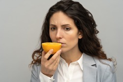 Sick business woman trying to sense smell of half fresh orange, has symptoms of Covid-19, corona virus infection - loss of smell and taste. One of the main signs of the disease. Studio grey background