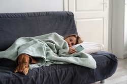Sad woman lying on sofa with dog, using smart phone in hands wait for boyfriend call or text, feels depressed. Thoughtful female think about relationships problems. Mental health, loneliness concept.