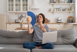Tired millennial woman suffers from stuffiness and an inoperative air conditioner, waving blue fan sitting on couch at home working on laptop computer. Overheating high temperature, hot summer weather