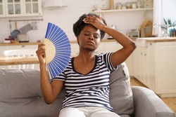 Tired African American woman suffering from heatstroke flat without air-conditioner, waving blue fan, sitting on sofa at home. Black girl cooling in hot summer weather. Overheating, high temperature.