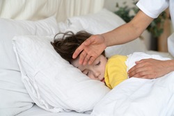 Caring mom check small sick boy temperature with hand. Ill kid with flu or covid illness sleep in bed under blanket with concerned mother on bedside. Child cure at home and parent healthcare concept