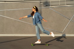 Young caucasian woman in trendy jeans clothes ride longboard skate over concrete wall in modern urban space or skatepark at sunset. Stylish curly brunette girl in trend glasses skating on skateboard