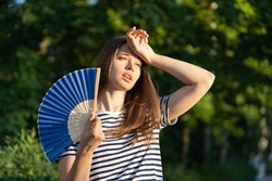 Young woman suffering heatstroke outdoors using fan to get fresh air. Unhappy girl feel bad of hot temperature outside. Frustrated female tired of summer season heat in fatigue touch forehead sweating