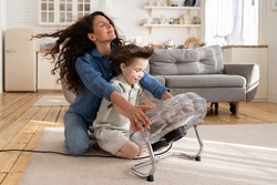 Cheerful young mom or nanny have fun with preschool boy at home sitting together in front of big fan dlowing cooling wind in living room. Carefree caring woman of 30 spend time with little son bonding