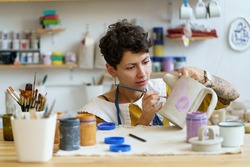 Young creative woman small business owner work in art studio with pottery for handmade shop. Girl relax with painting, ceramics after work create craft kitchenware in workshop. Artistic hobby concept